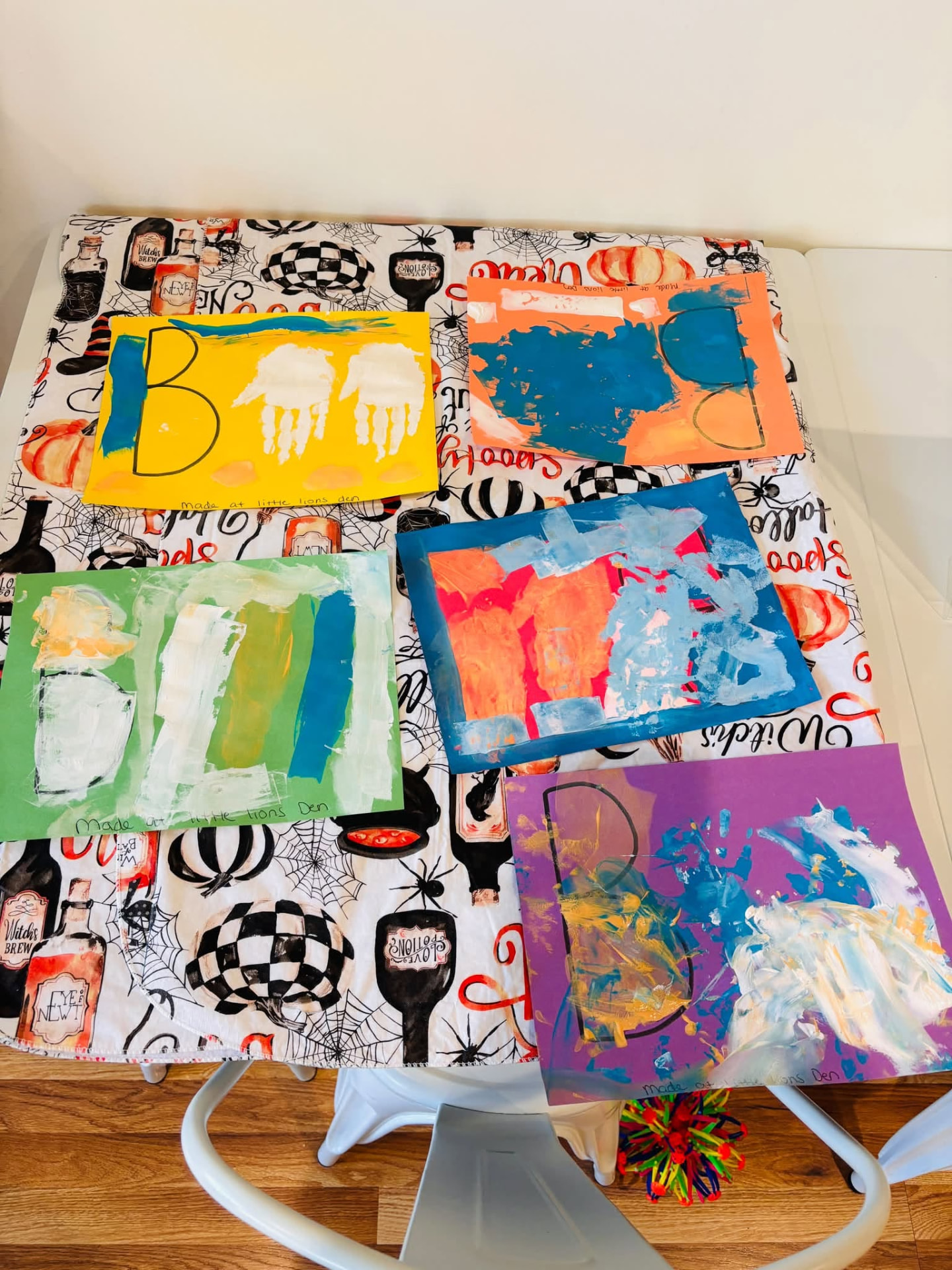 Five finger-painted children's crafts spelling BOO with handprint ghosts, drying on a Halloween table runner at Little Lions Den